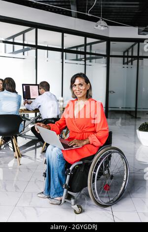 Latin transgender woman sitting in wheelchair in modern office using laptop with coworkers in Mexico City Stock Photo