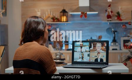 Woman on video call with workmates at home waiting for christmas eve holiday celebration. Young adult sitting in kitchen with ornaments and decorations using laptop to talk to friends Stock Photo