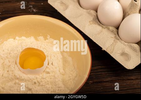 A broken white chicken egg in a bowl of wheat flour and several whole eggs in a tray made of white cardboard, close-up selective focus Stock Photo