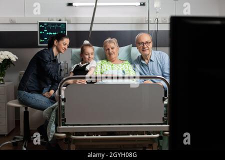 Cheerful family spending time with sick grandmother watching movie on tv during medical recovery visiting her in hospital ward. Senior woman patient resting in bed waiting for healthcare treatment Stock Photo