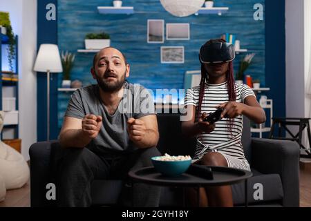 Interracial couple using vr glasses and controllers for fun at home. Mixed race young people with technology playing video game with goggles, headset and console on television. Stock Photo