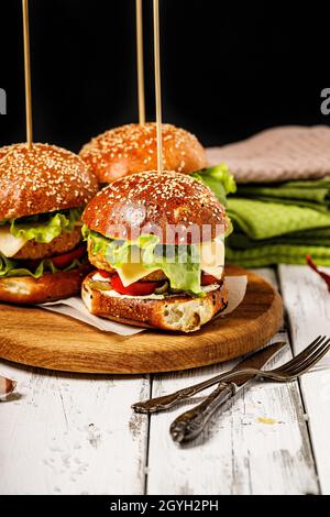 Appetizing homemade chicken burger with cheese, fresh salad and vegetables on a round cutting board. Lettuce leaves, tomatoes and onions. Traditional Stock Photo