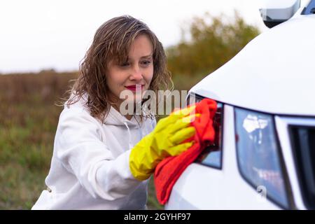 A pretty young woman in a white hoodie and yellow rubber gloves wipes the headlights of a car with a bright red rag on a warm autumn day. Selective fo Stock Photo