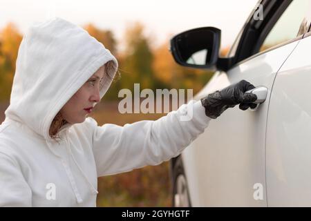A beautiful young woman in a white sweater with a hood and black gloves opens the car door to steal it. Selective focus. Close-up. Portrait Stock Photo
