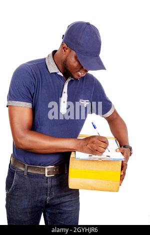 a delivery man standing in cap on white background holding a cardboard box writing on a notepad smiling. Stock Photo