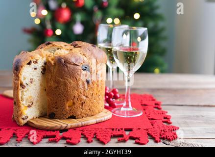 Panettone and white wine glasses on a wooden table with Christmas decorations and tree in the background. Copy space. Stock Photo