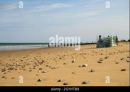 A couple sitting behind a colourful plastic sheet shielding them from the wind on Mundesley beach, Norfolk, England. Stock Photo