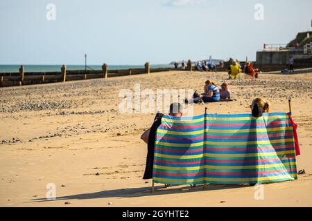 A couple sitting behind a colourful plastic sheet shielding them from the wind on Mundesley beach, Norfolk, England. Stock Photo
