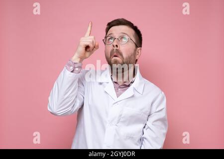 Surprised doctor in a white coat and glasses points his finger up and looks puzzled. Pink background. Stock Photo
