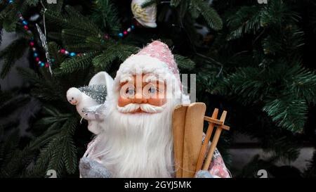 Toy Santa Claus with a bag of gifts on his shoulder and wooden skis. Doll Santa Claus with glasses and a white beard near the Christmas tree. Stock Photo