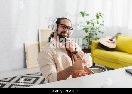 dreamy and smiling hispanic man with smartphone listening music in headphones at home Stock Photo