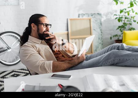 thoughtful latin designer looking at digital tablet while sitting with legs on desk near blurred smartphone Stock Photo