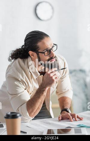 pensive hispanic designer holding pen and looking away while working at home Stock Photo