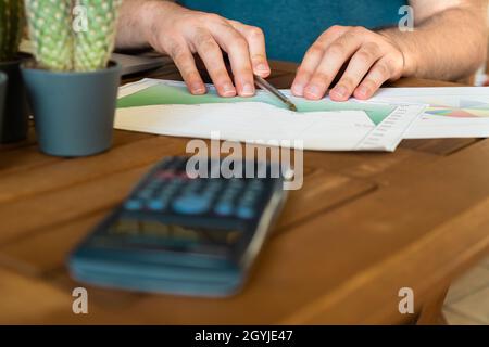 Unrecognized young man analyzing charts on desk with a calculator while working from bright home office. Stock Photo