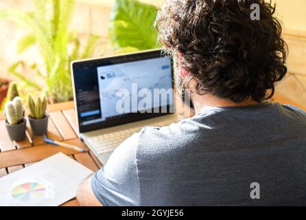 Young man sitting at table investing in stock market with laptop during work remotely from home office. Stock Photo
