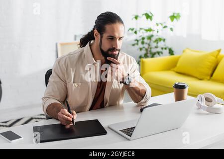 thoughtful hispanic retoucher touching beard while working with laptop and graphic tablet at home Stock Photo