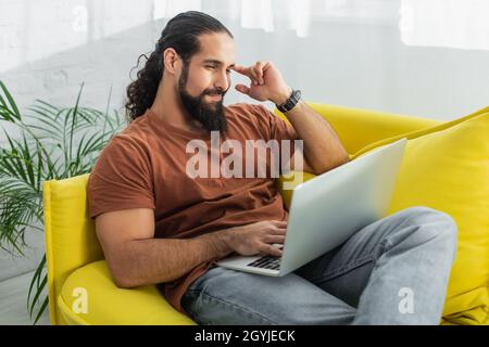 positive and thoughtful hispanic man looking at laptop while sitting on sofa at home Stock Photo