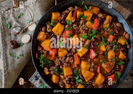 Stew with ground beef, pumpkin, kidney beans, bell peppers and tomatoes. Healthy autumn and winter meal served in a cast iron skillet on wooden table. Stock Photo