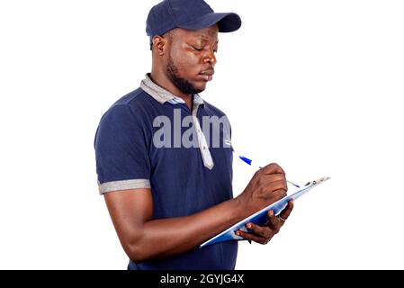 young delivery man standing in blue uniform on white background jotting down his orders on a notepad. Stock Photo