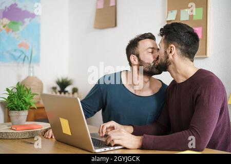 Happy gay couple kissing while using laptop in living room at home - LGBT love and technology concept Stock Photo