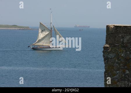 Gaff rigged wooden sailing boat setting sail with tanker in the background, Cornwall Stock Photo