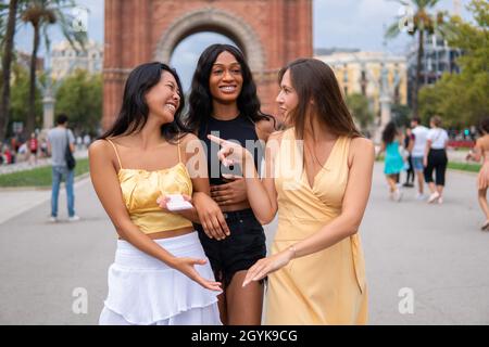 Surprised young woman pointing at laughing girlfriend while chatting and discussing news during walk on city street in summer day Stock Photo