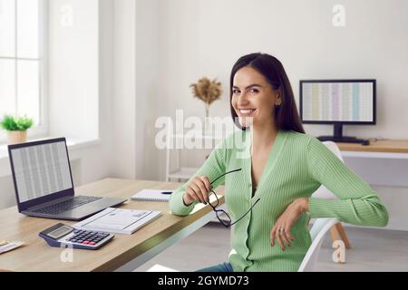 Portrait of beautiful happy female accountant sitting at her workplace in office. Stock Photo