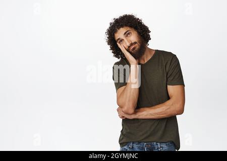 Sad and bored curly guy lean on hand, staring reluctant and unamused, standing in casual clothes over white background Stock Photo