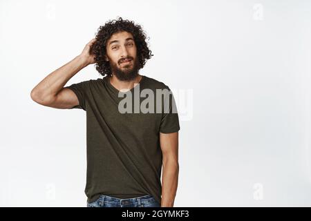 Clueless young man looking unsure, scratching his head indecisive, standing in t-shirt over white background Stock Photo