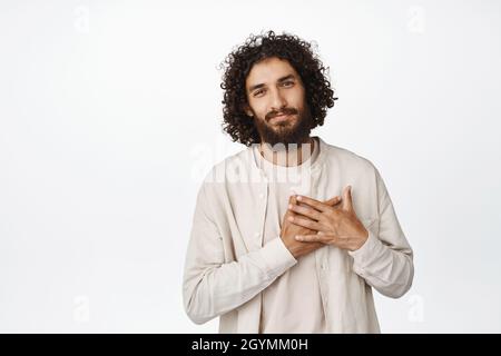 Image of young middle aged man holding hands on heart, looking caring and tender at camera, express gratitude, thankful, standing over white Stock Photo