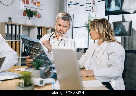 Two focused medical workers using modern gadgets while examining results of x ray scan. Group of scientists practitioners gathering at conference room for discussing ways of patient treatment. Stock Photo