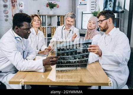 Five qualified mixed race doctors in uniform sitting at desk and looking at x ray scan. Group of therapists with one colleague who using wheelchair discussing ways of patient treatment. Stock Photo