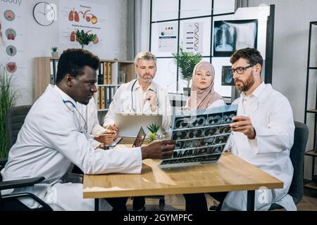 Five qualified multiethnic doctors in uniform sitting at desk and looking at x ray scan. Group of therapists with one colleague who using wheelchair discussing ways of patient treatment. Stock Photo