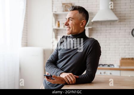 Handsome smiling man using mobile phone while leaning on table at home kitchen Stock Photo