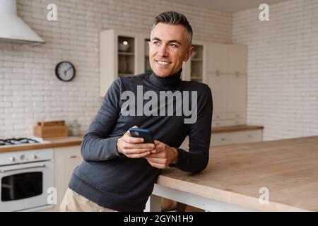 Handsome smiling man using mobile phone while leaning on table at home kitchen Stock Photo
