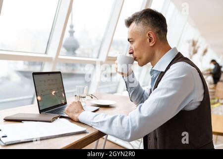 Confident pensive handsome businessman in smart clothes working on laptop computer while sitting in the cafe indoors Stock Photo