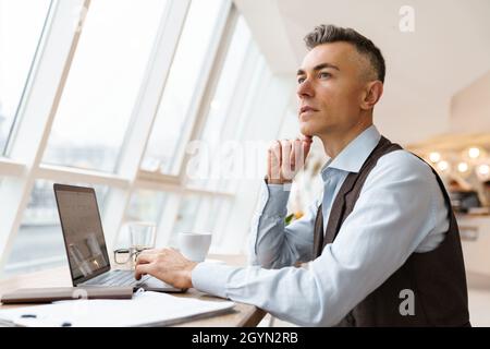Confident pensive handsome businessman in smart clothes working on laptop computer while sitting in the cafe indoors Stock Photo