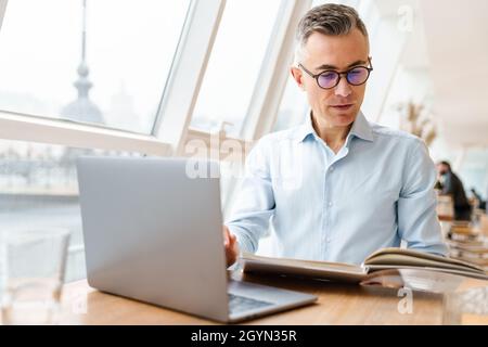 Confident handsome businessman in smart clothes working on laptop computer while sitting in the cafe indoors, looking through documents Stock Photo