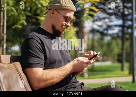 Side view of blogger man wear casual clothes sit on bench while surfing smartphone outdoor in street Stock Photo