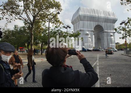 Person uses a smartphone to photograph the Arc de Triomphe wrapped in silver-blue fabric fastened with red ropes in the Place Charles de Gaulle in Paris, France. The Arc de Triomphe was wrapped for two weeks being converted to an artwork as it was designed by Christo and Jeanne-Claude in September 2021. Stock Photo