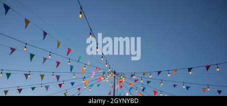 Garland with colorful pennants and light bulbs on pole over blue sky. Long format Stock Photo