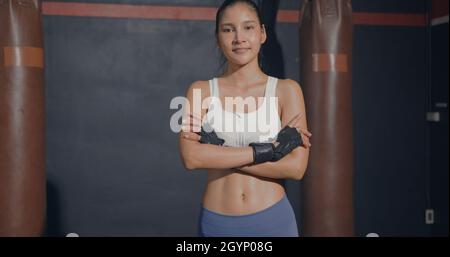 Asian woman boxer standing inside a boxing gym fitness club studio with her hands folded, Woman boxing hand wraps crossed arms she looking to camera, Stock Photo