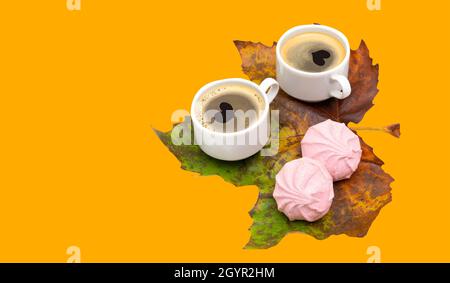 Two coffee cups with heart shapes on the foam and rose marshmallows on a large maple leaf isolated on a yellow background. Romantic fall still life. Stock Photo