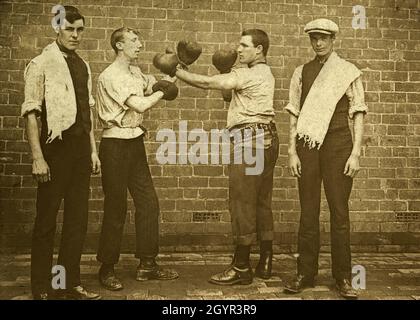 Early 1900s boxers in sparring pose, flanked by their seconds with towels draped over their shoulders. The boxer on the right appears to be wearing a championship belt. Vintage photograph.  A semi-legible hand-written note handwritten on the back of the original card mount indicates the photograph may have been taken in Newport in Wales. Stock Photo