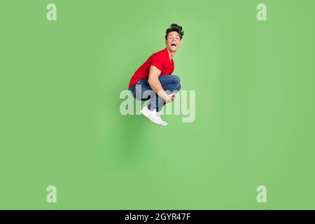 Photo of impressed funny young guy dressed red t-shirt jumping high smiling isolated green color background Stock Photo