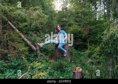 Woman resting in forest in park dressed in denim suit and boots. Young caucasian woman sitting on tree stump surrounded by forest after hiking Stock Photo