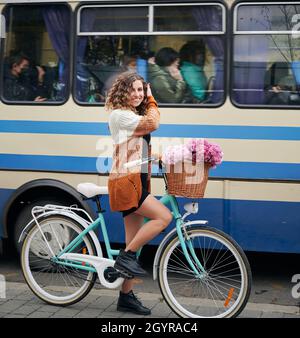 Close up of smiling young woman with curly hair riding bicycle with bouquet pink fresh flowers on blue and white bus background. Concept of walking with bicycle in cetre city. Stock Photo