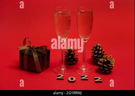A composition of New year object, wooden 2022 numerals, golden pine cones, Christmas gift in glitter wrapping paper with golden bow, champagne flutes Stock Photo