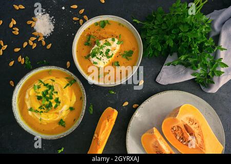 Homemade roasted squash cream soup with parsley on the top on rustic bowls and raw squash pieces and seeds on black kitchen table Stock Photo