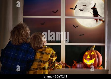 Kids watch witch on Halloween night. Children sitting at window watching bats and ghosts by full moon. Trick or treat fun. Boy dress up. Home pumpkin Stock Photo